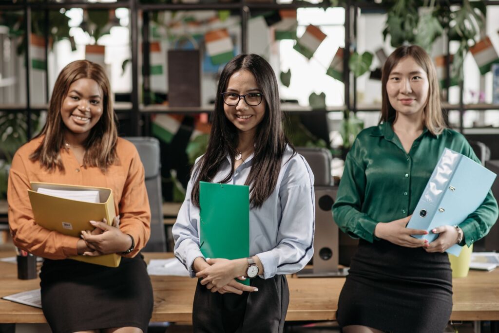 Three diverse women smiling and holding folders in a modern office environment.