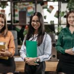 Three diverse women smiling and holding folders in a modern office environment.