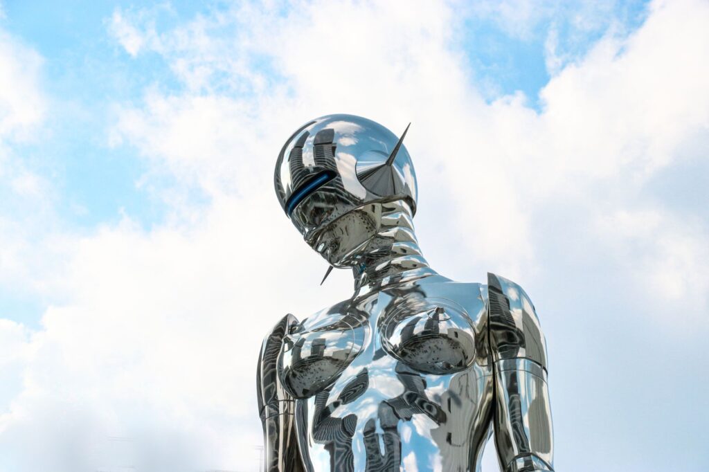 A sleek chrome robot sculpture stands against a bright blue sky background.
