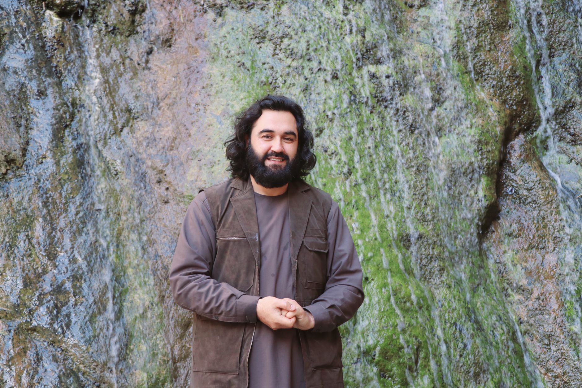 A bearded man in a brown vest stands by a mossy waterfall.