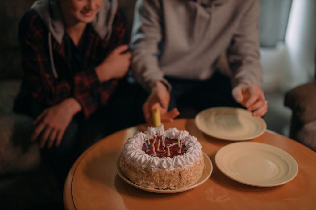 Close-up of a birthday cake with lit candles and two friends celebrating indoors.