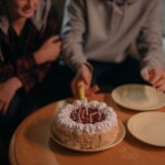 Close-up of a birthday cake with lit candles and two friends celebrating indoors.