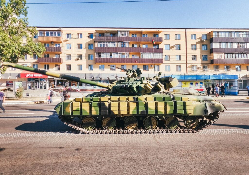 Camouflaged military tank parked in front of an urban residential building during a sunny day.