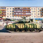 Camouflaged military tank parked in front of an urban residential building during a sunny day.