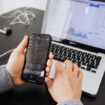 Person analyzes stock data with a smartphone and laptop, indoors.